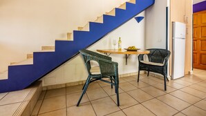 Una escalera con peldaños azules que lleva a una plataforma, dos sillas de mimbre, una mesita redonda con una botella y un vaso, y un refrigerador al fondo.