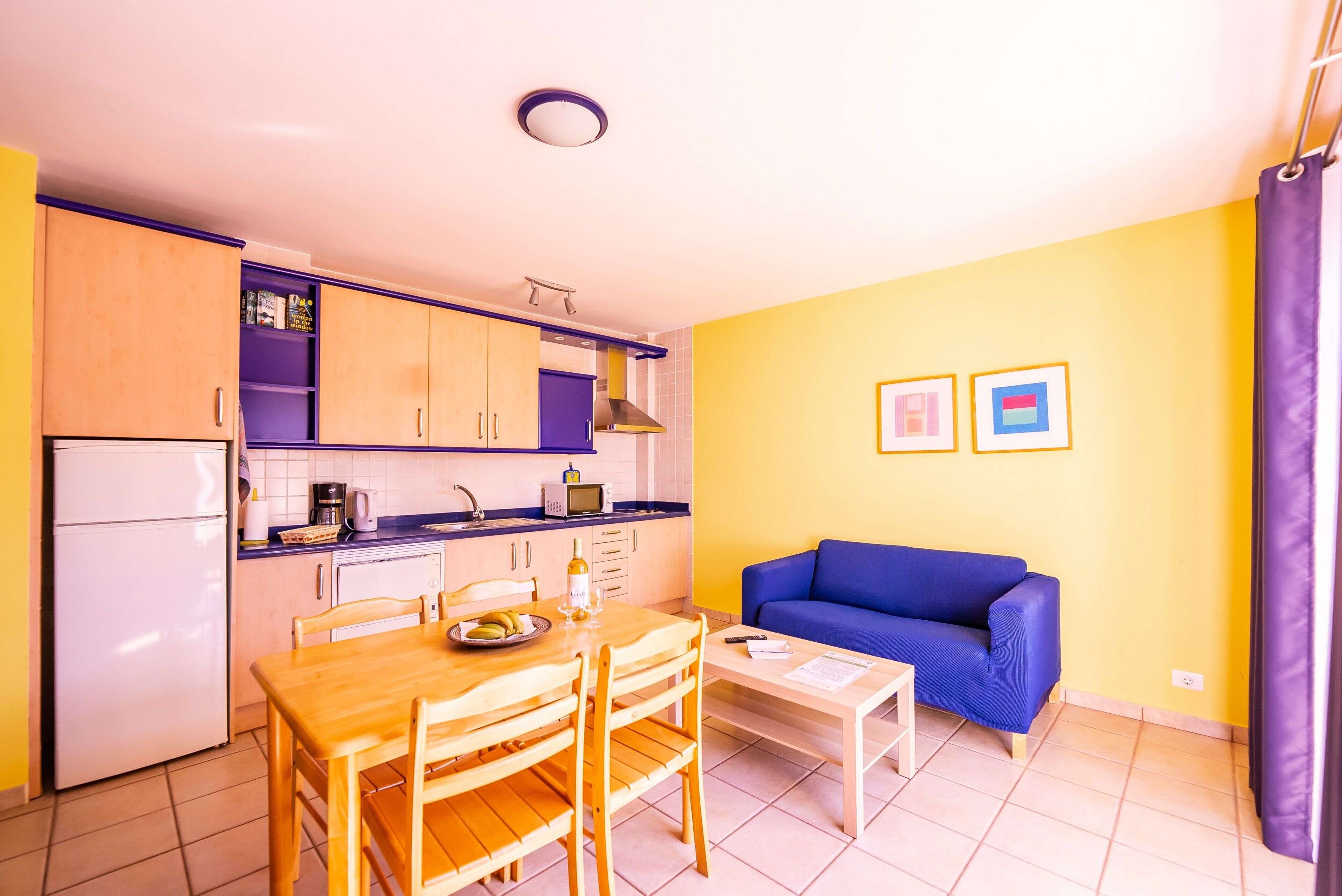 A compact kitchen with a dining table and chairs, a blue sofa, and a small table with a remote control and papers.