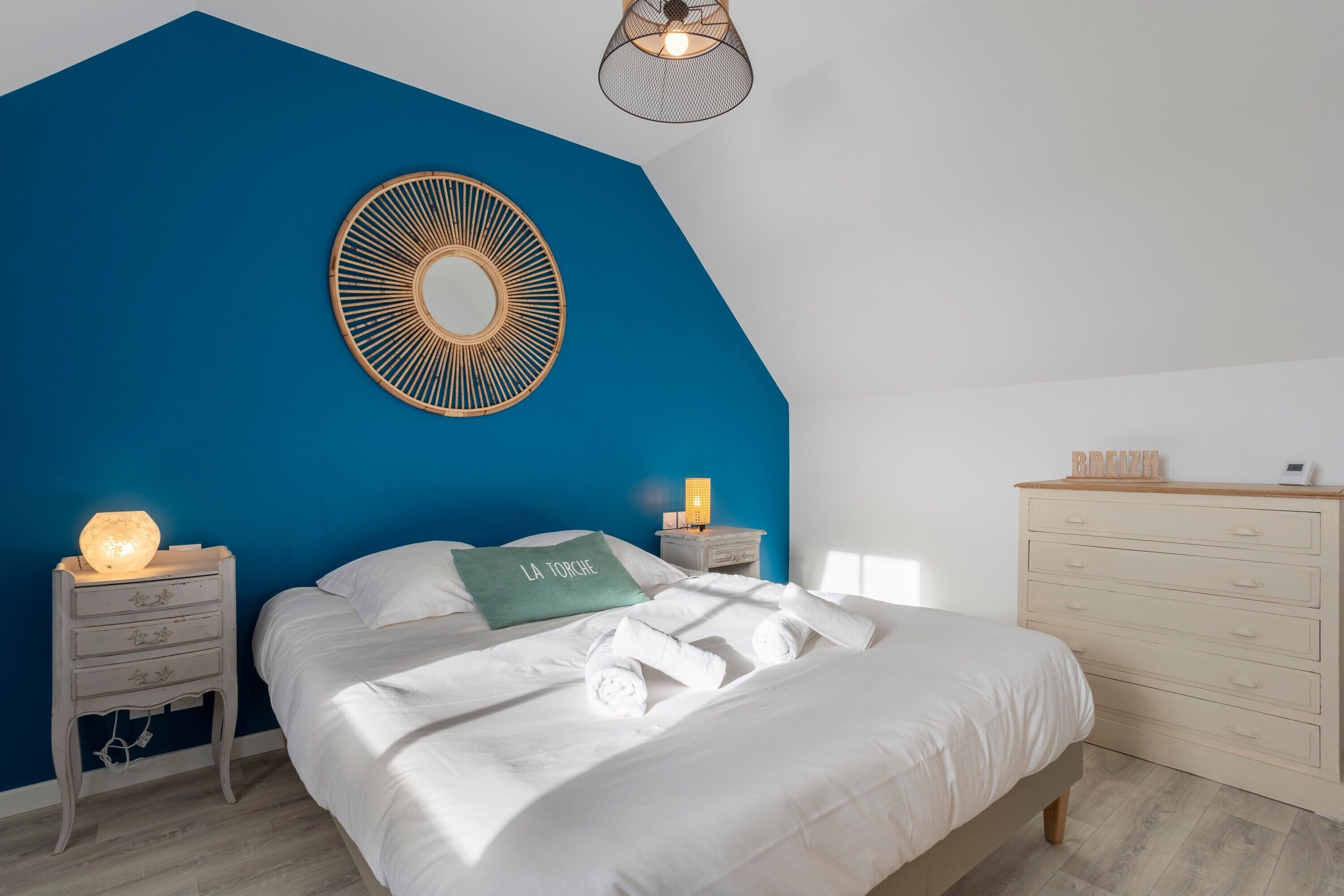 Une chambre avec un mur d’accent bleu, un lit avec du linge de lit blanc, une table de chevet avec une lampe, une commode et un miroir rond fixé au mur.