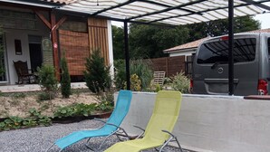 A covered outdoor seating area with two lounge chairs, a vehicle, and a house with a tiled roof.