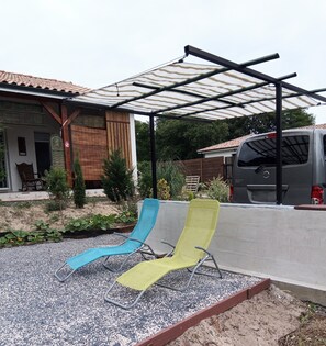 A covered outdoor seating area with two lounge chairs, a vehicle, and a house with a tiled roof.