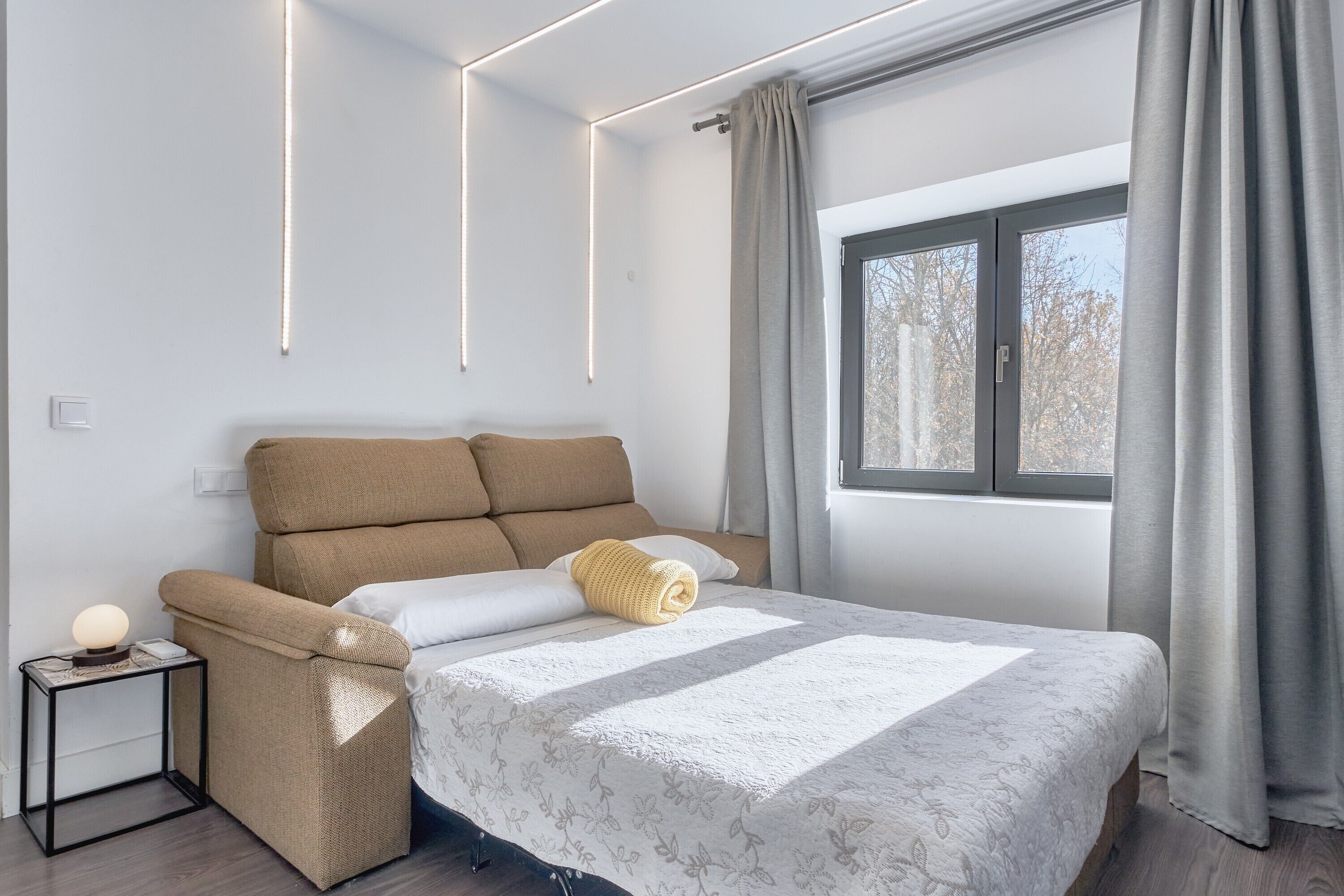 Camera da letto moderna con divano letto, comodino con lampada, finestra con tende e vista su alberi.