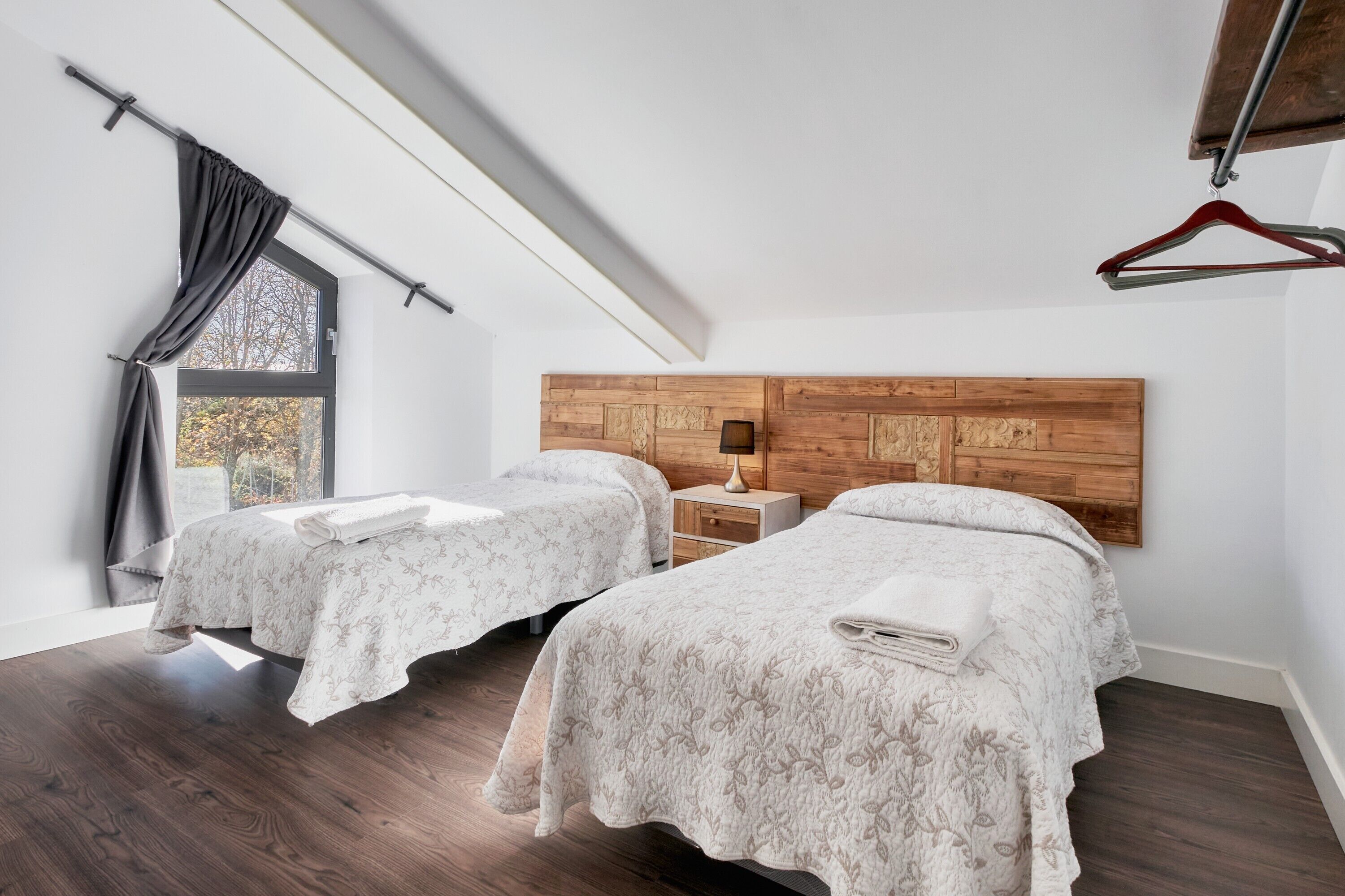 Une chambre avec deux lits, une tête de lit en bois, une table de chevet, une lampe suspendue et une fenêtre avec des rideaux.