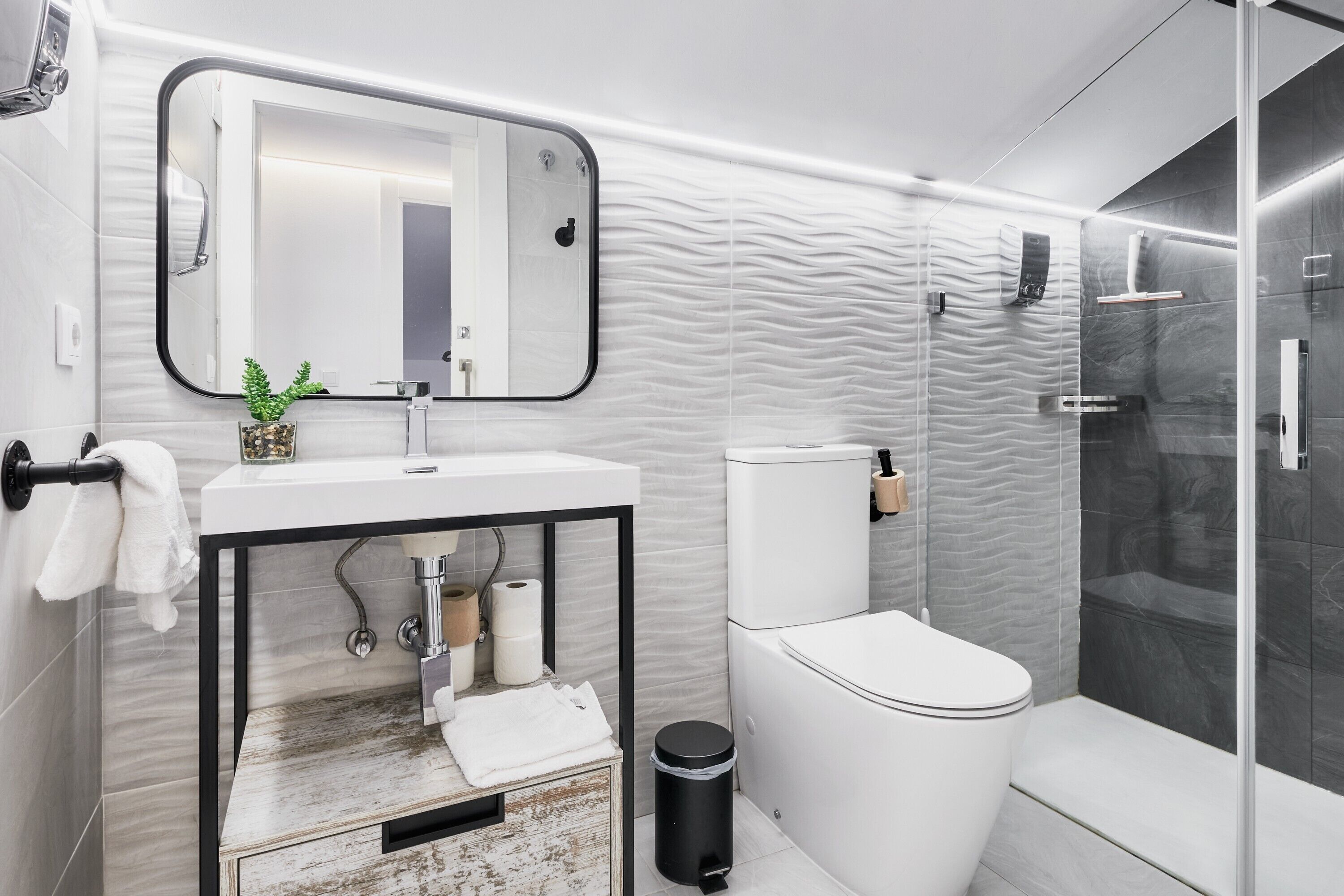 A modern bathroom with a white sink, mirror, and toilet, a glass shower enclosure, and a textured wall design.