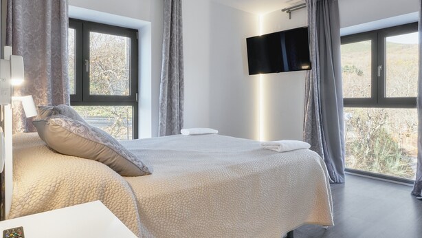 A bedroom with a large bed, a flat-screen TV mounted on the wall, and a window with curtains.