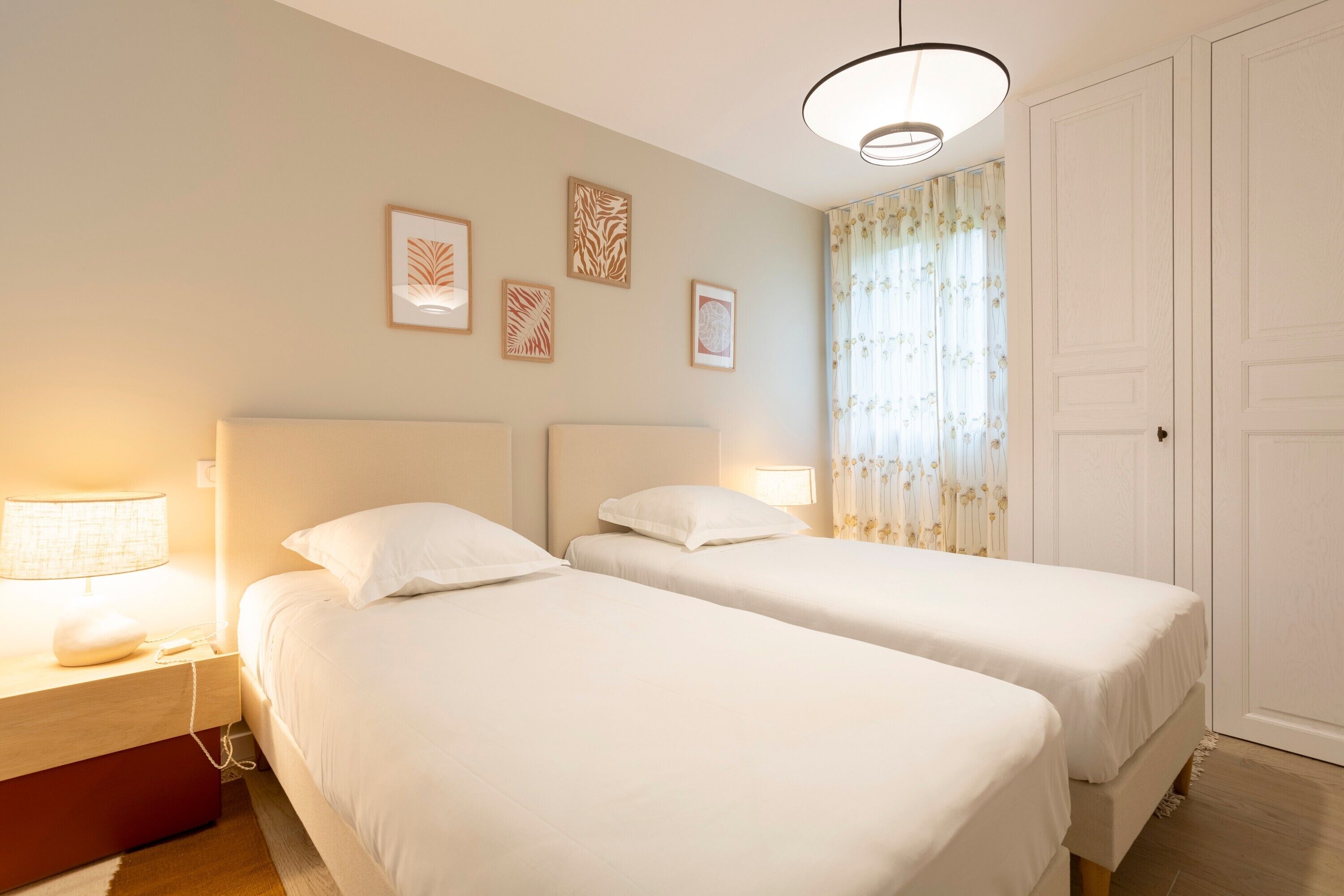 Une chambre d’hôtel avec deux lits, une table de chevet avec une lampe, une porte et des œuvres d’art encadrées au mur.