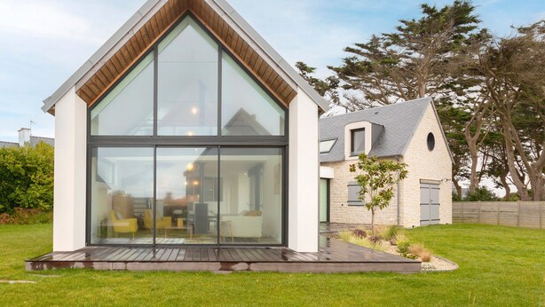 Una casa moderna con un amplio ventanal de vidrio, una terraza de madera y fachada de piedra.