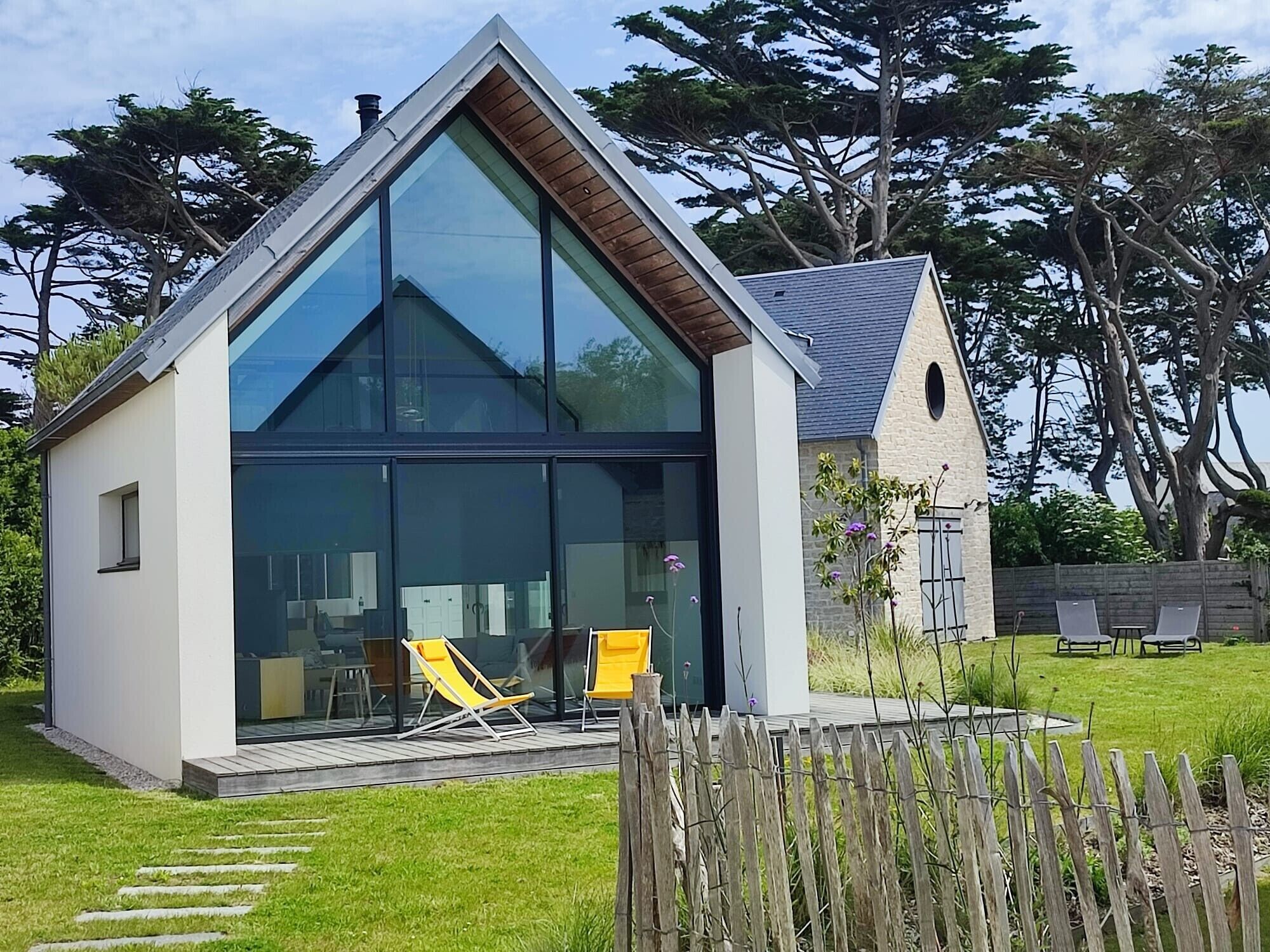 Une maison moderne dotée d’une grande façade vitrée, d’une terrasse en bois et d’un petit jardin.