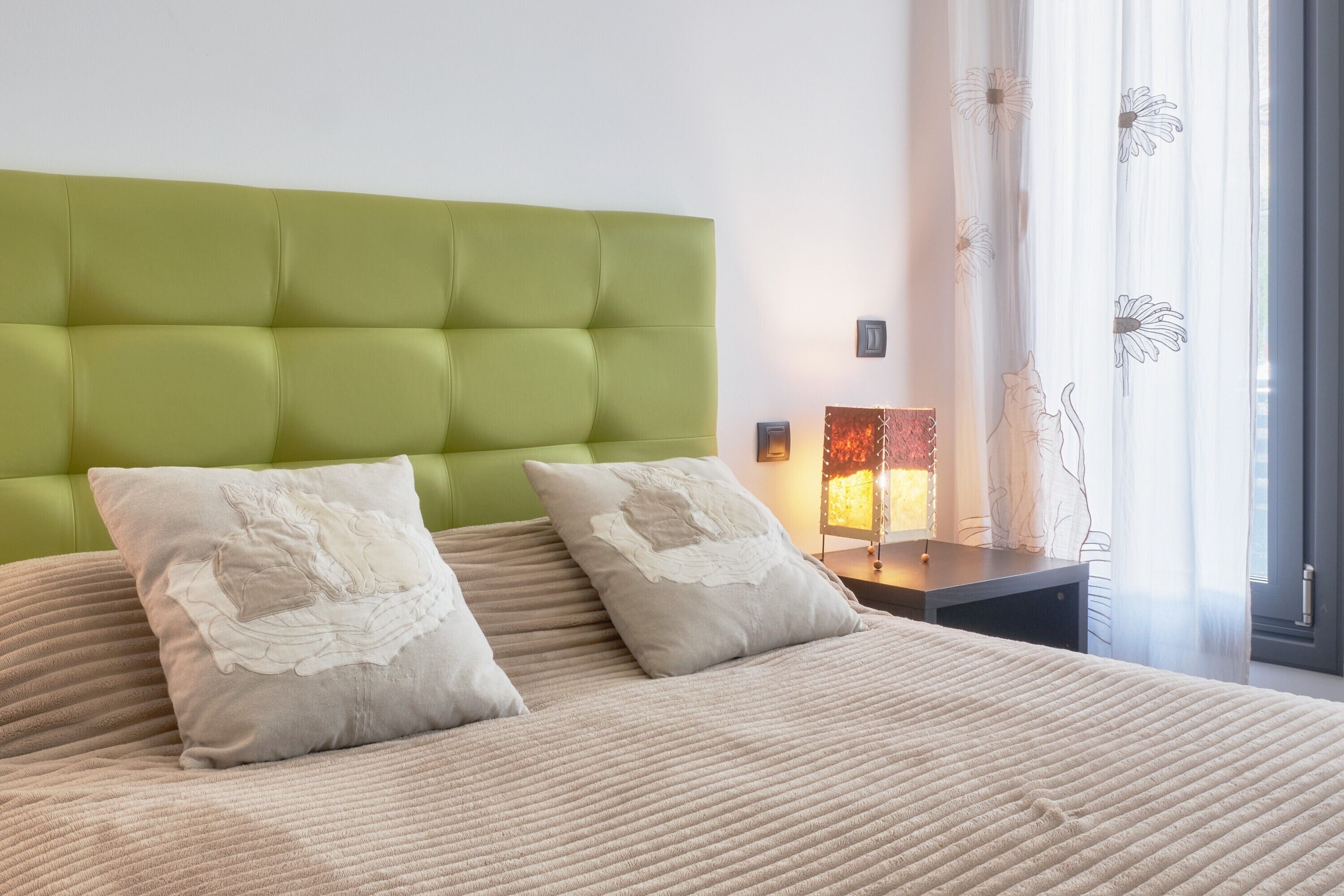 A bed with a green upholstered headboard, two pillows with floral embroidery, a bedside table with a lamp, and a window with sheer curtains.