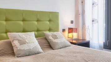 A bed with a green upholstered headboard, two pillows with floral embroidery, a bedside table with a lamp, and a window with sheer curtains.