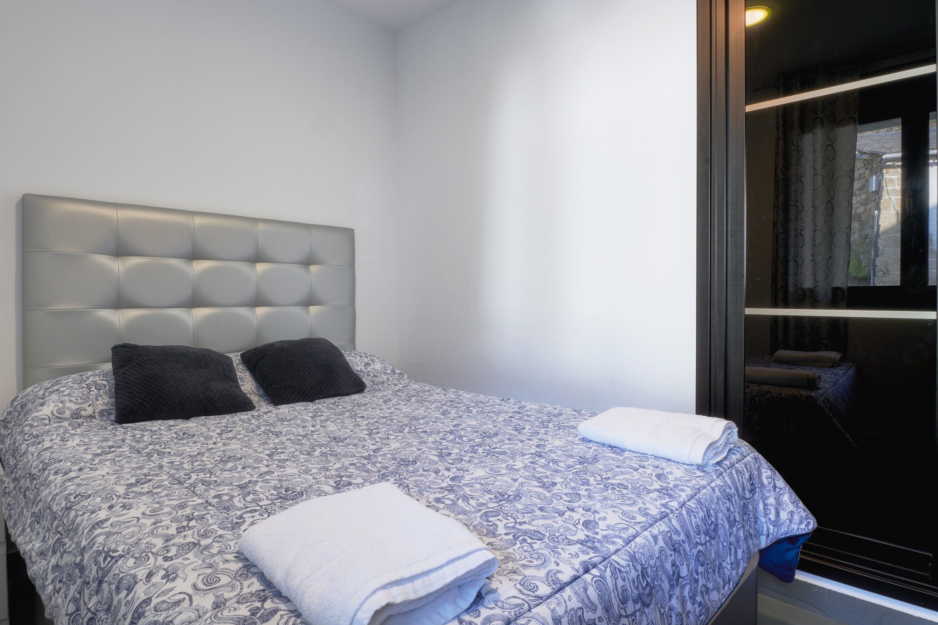 Un dormitorio con cabecera capitonada, dos almohadas negras, una colcha estampada y una toalla blanca doblada con cuidado.