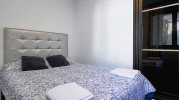 Un dormitorio con cabecera capitonada, dos almohadas negras, una colcha estampada y una toalla blanca doblada con cuidado.
