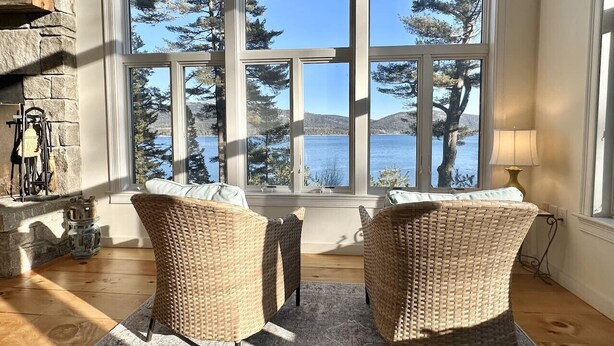 Une pièce avec deux fauteuils en osier, une cheminée en pierre et une grande fenêtre donnant sur un plan d’eau et des arbres.