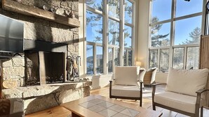 Une cheminée en pierre, deux canapés blancs et une table basse en bois dans une pièce bien éclairée, dotée de grandes fenêtres.