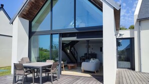 Une maison moderne dotée d’une grande façade en verre, d’une terrasse avec mobilier d’extérieur et d’un escalier apparent menant à un étage supérieur.