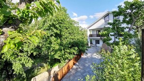 A modern two-story house with a balcony, surrounded by lush greenery and a stone wall.