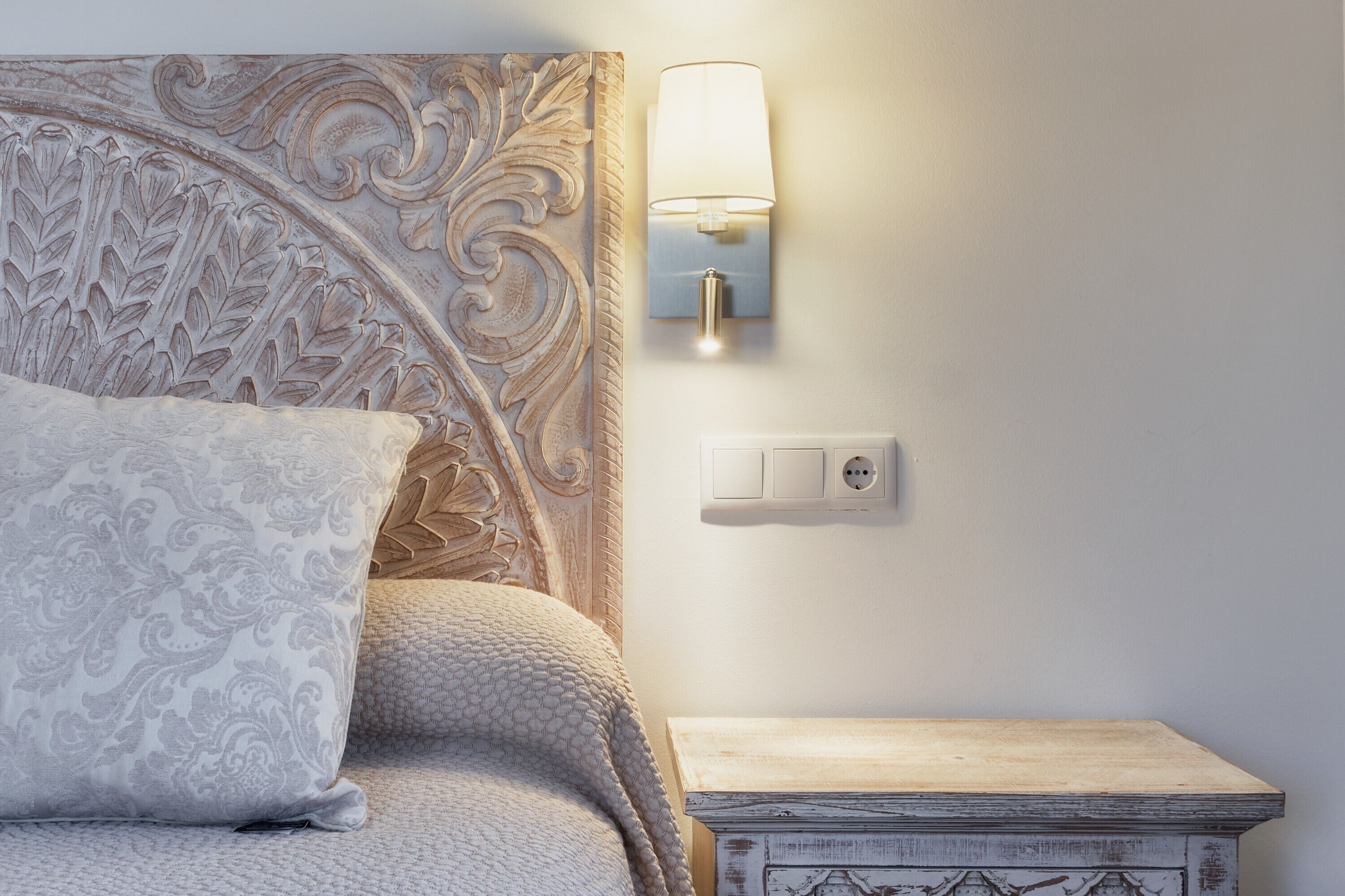 Una cama con un coj ín estampado, una lámpara fijada a la pared y una mesita de noche de madera.