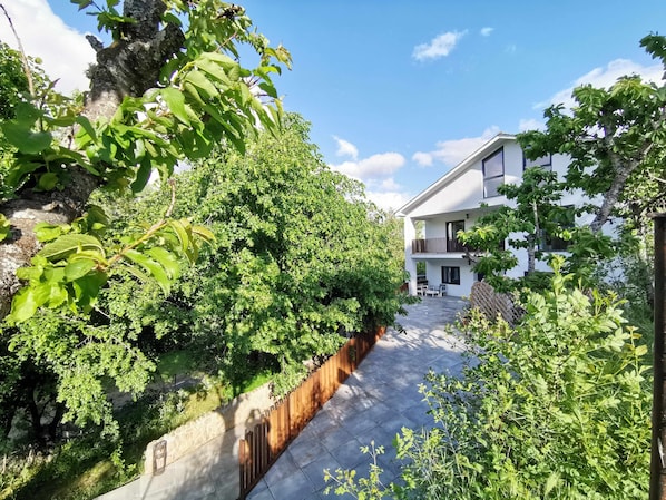 A modern two-story house with a balcony, surrounded by lush greenery and a stone wall.