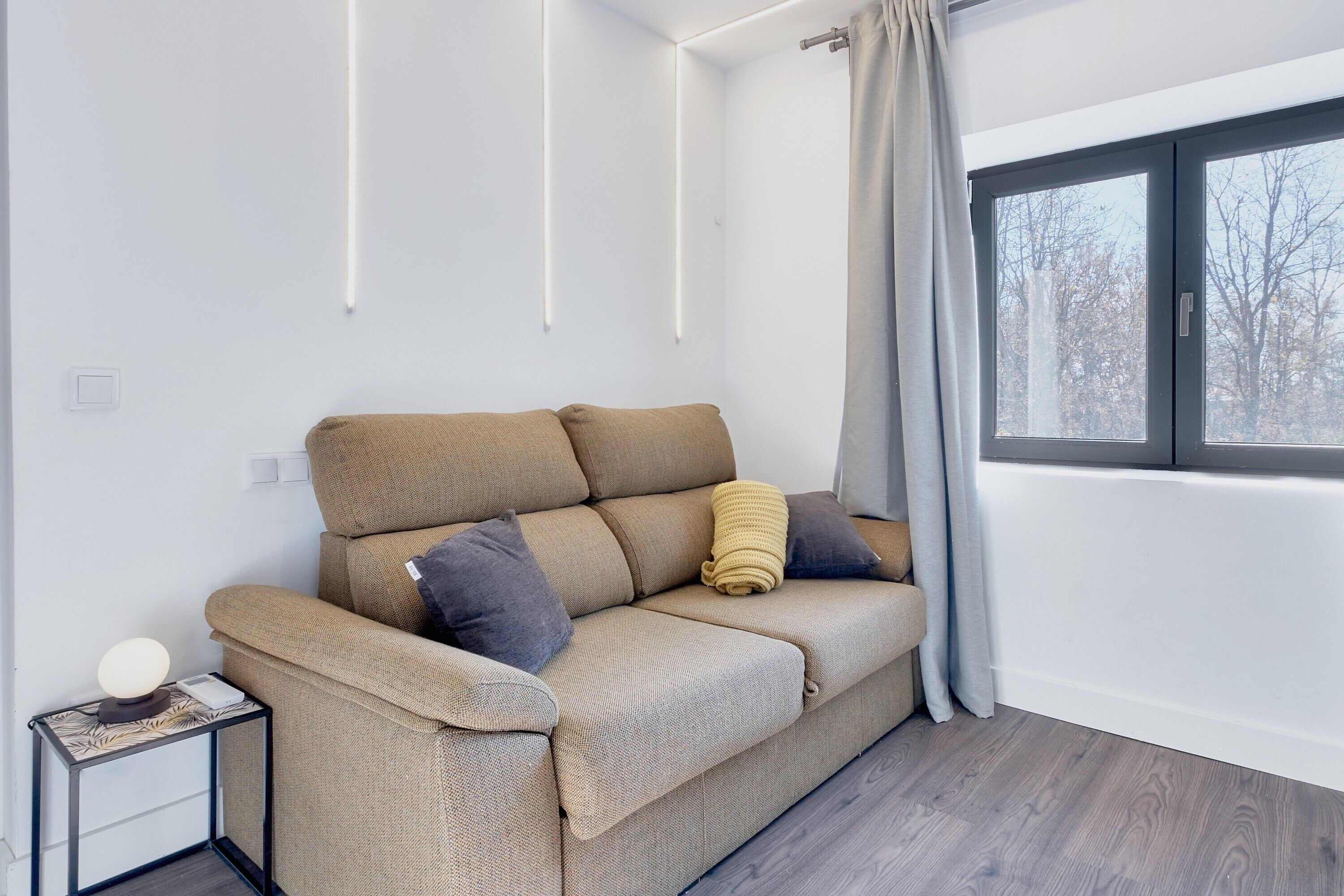A modern living room with a beige sofa, a side table with a lamp, and a window with curtains.