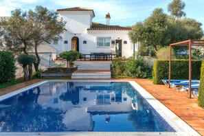 Vue sur une maison avec piscine, prise depuis le bord de la piscine, montrant une terrasse avec des chaises et un jardin.