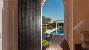 Visão através de uma porta arqueada para uma área de piscina com mesa e cadeiras, uma planta em vaso e um abajur.