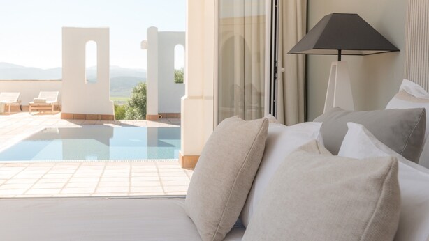 A bedroom with a bed, pillows, a lamp, and a view of a pool and outdoor seating.