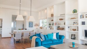 A spacious living room with a blue sofa, white dining area, and a bookshelf filled with decorative items.