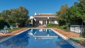 Una piscina con superficie azul cristalina, un edificio blanco con arcos y un jardín con sillas azules.