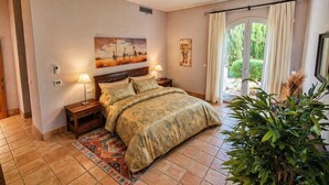 Un dormitorio con cama, dos mesitas de noche, una lámpara de noche, una ventana con cortinas, un cuadro en la pared y una planta en maceta.