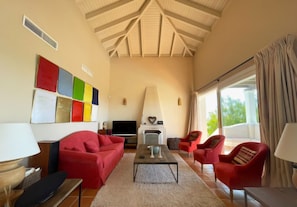 Una sala de estar con techo alto, chimenea, televisor, sofás rojos y un ventanal con cortinas.