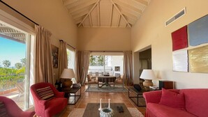 Ein geräumiges Wohnzimmer mit hoher Decke, ein Balkon mit Vorhängen, ein Essbereich mit Tisch und Stühlen und ein rotes Sofa.