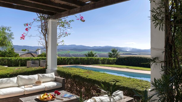 Ein Poolbereich mit einer Couch, einem Holztisch mit Früchten und Blick auf Berge.