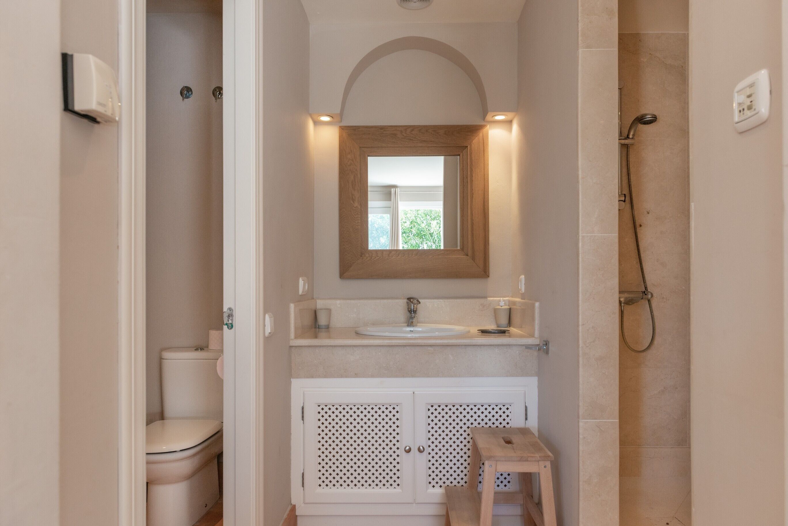 Ein Badezimmer mit Toilette, Waschbecken, Spiegel, Dusche und einem kleinen hölzernen Hocker.