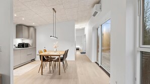 Une cuisine moderne avec un coin repas, comprenant une table et des chaises en bois, un luminaire suspendu et une vue sur l’extérieur grâce à une grande fenêtre.