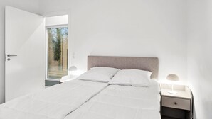 Une chambre à coucher avec un lit, deux tables de chevet avec des lampes, une porte donnant sur un balcon et une vue sur les arbres à l’extérieur.