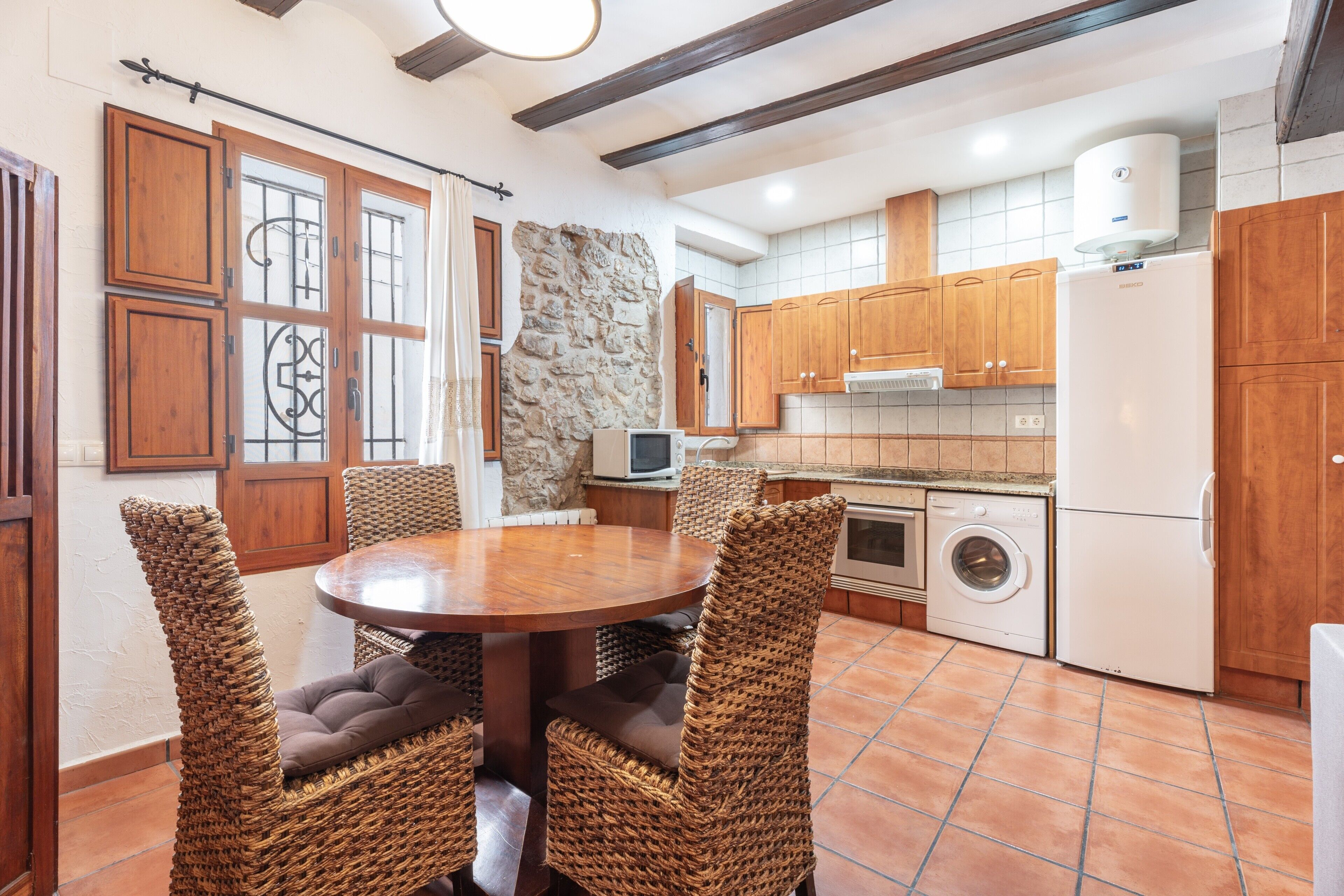 A kitchen with a stone wall, wooden cabinets, a washing machine, and a dining area with wicker chairs.