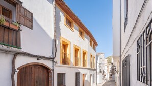A narrow street with white buildings, wooden doors, and balconies with plants.