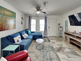 Una sala de estar moderna con un sofá azul, un sillón rojo, una mesita redonda y un televisor sobre un mueble de madera.