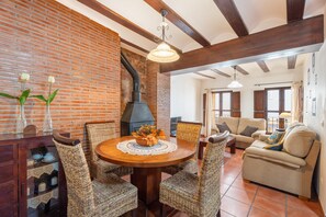A dining area with a brick wall, a fireplace, wicker chairs, a round wooden table, and a sofa in the background.