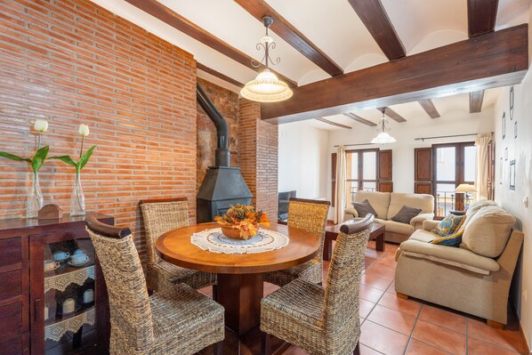 Un comedor con pared de ladrillo, chimenea, sillas de mimbre, una mesa redonda de madera y un sofá al fondo.