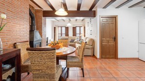 Un comedor con pared de ladrillo, vigas de madera, sillas de mimbre, una mesa redonda y una chimenea.