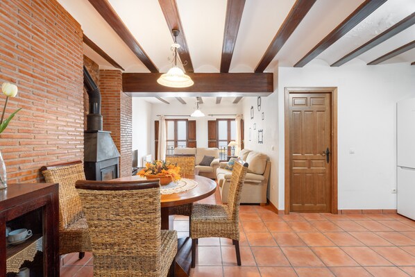 A dining area with a brick wall, wooden beams, wicker chairs, a round table, and a fireplace.
