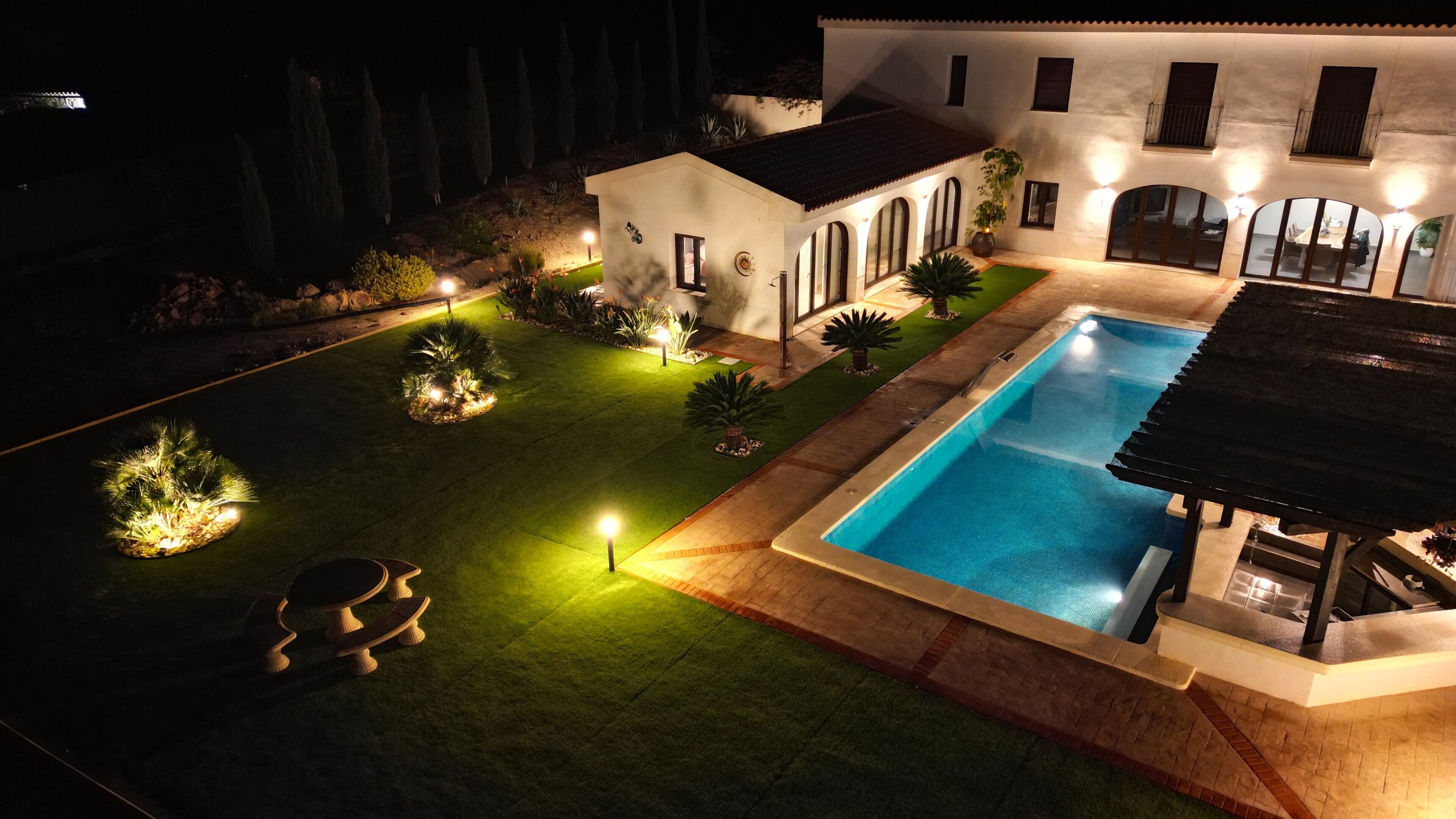 Una propiedad residencial bien iluminada con piscina, un jardín ajardinado y una casa con múltiples ventanas y un balcón.