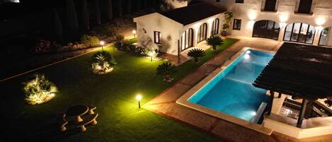 Una propiedad residencial bien iluminada con piscina, un jardín ajardinado y una casa con múltiples ventanas y un balcón.