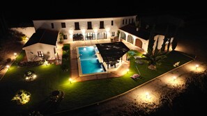 Een groot huis met zwembad, terras en een goed verlichte tuin 's nachts.