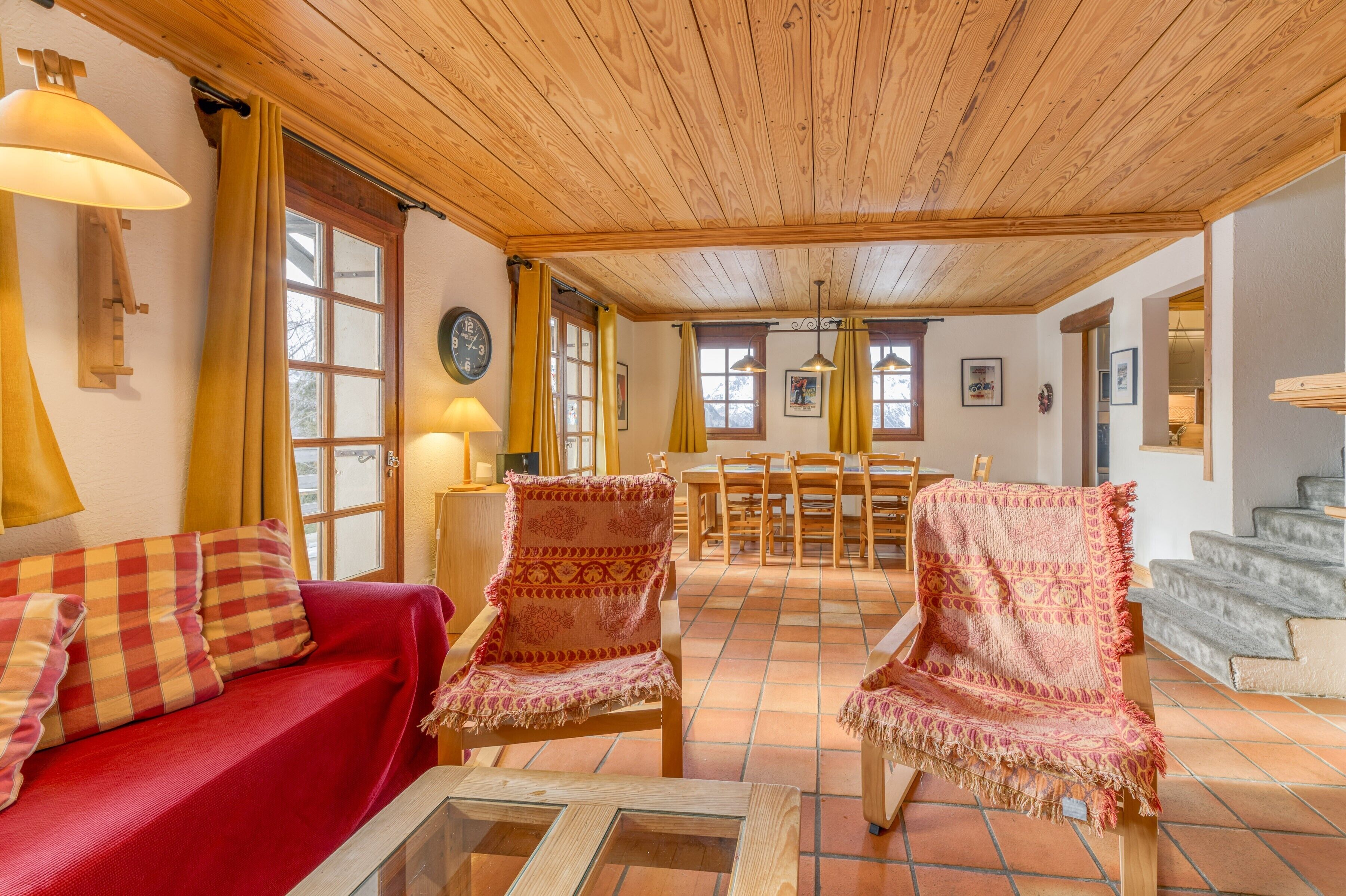 Un salon chaleureux avec un canapé rouge, des fauteuils à motifs, un plafond en bois et un sol carrelé.