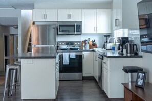 A modern kitchen with white cabinets, stainless steel appliances, and a central island with stools. - The Spruce Willis Suite (Grande Prairie)