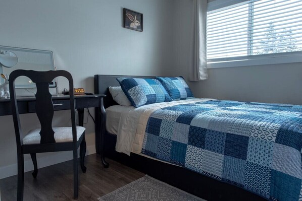 A bedroom with a bed, a desk, a chair, a clock, and a window with blinds. - The Spruce Willis Suite (Grande Prairie)