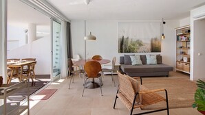 A modern living room with a sofa, dining table, chairs, and a large window. - Apartment 'Golden Sky' with Shared Pool, Wi-Fi and Air Conditioning (Cabanas de Tavira)