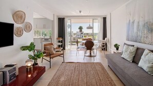 A modern living room with a sofa, coffee table, and TV. There is a dining area with chairs and a table. The room has large windows and a view of the outdoors. - Apartment 'Golden Sky' with Shared Pool, Wi-Fi and Air Conditioning (Cabanas de Tavira)